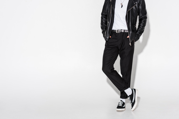 cropped view of man in leather jacket standing with crossed legs on white  - Foto, afbeelding