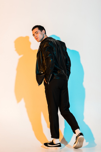 handsome man in black leather jacket standing with hand in pocket and looking at camera on white with illumination  - Φωτογραφία, εικόνα