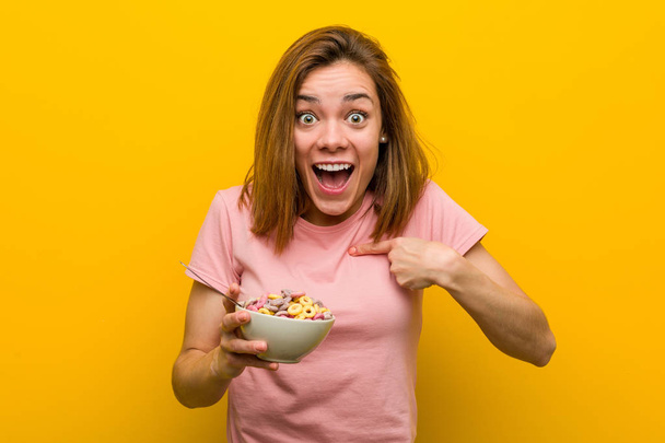 Mujer joven sosteniendo un tazón de cereales sorprendida señalándose a sí misma, sonriendo ampliamente
. - Foto, imagen