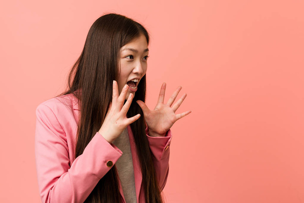 młody biznes chiński kobieta noszenie różowy garnitur krzyczy głośno, trzyma oczy otwarte i ręce napięte. - Zdjęcie, obraz