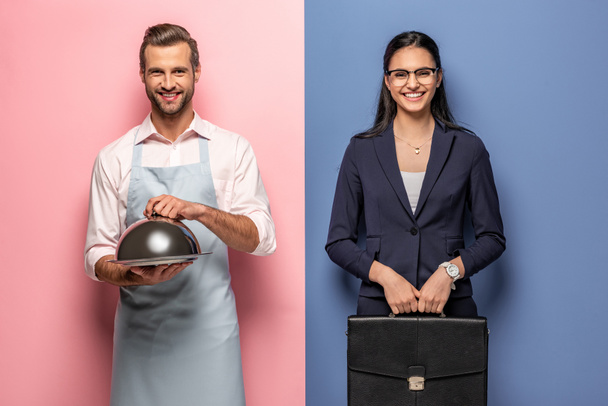 homme heureux dans tablier avec plateau de service et femme d'affaires avec mallette sur bleu et rose
 - Photo, image