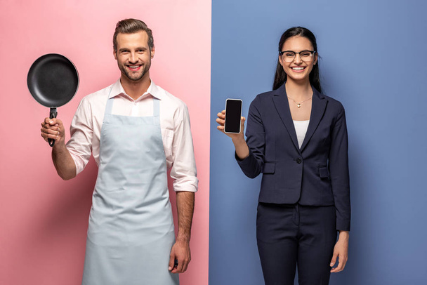 мужчина в фартуке держит сковородку в то время как деловая женщина держит смартфон с чистым экраном на голубом и розовом
 - Фото, изображение