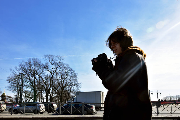 Mladý fotograf natáčí na jaře auta na ulicích s kamerou, na modrém pozadí, Sain-Petersburg, Rusko. Fotografie proti slunci, zadní světlo, boční světlo - Fotografie, Obrázek