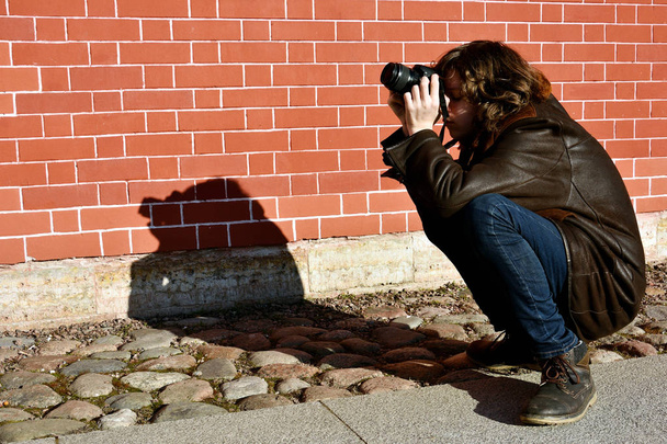 Joven fotógrafo (adolescente) se sentó con una cámara en sus manos para pensar en una imagen, luz solar directa, sombra oscura, fondo de pared de ladrillo
 - Foto, imagen