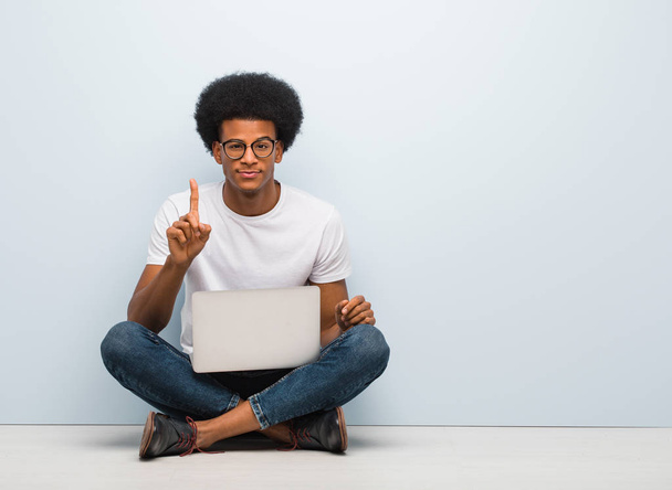 Młody czarny mężczyzna siedzi na podłodze z laptopem i pokazuje numer jeden - Zdjęcie, obraz