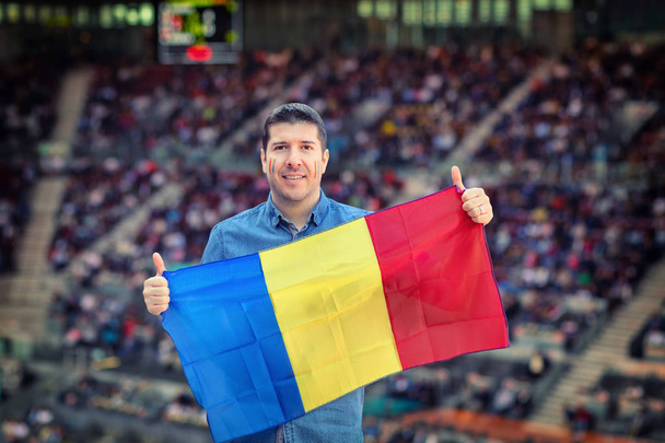 Happy caucasian man holding Romanian national flag in hands at international sport event, supporter at stadium cheering his favourite tennis player with blurred people in background  loyal fan in soccer arena - Photo, Image