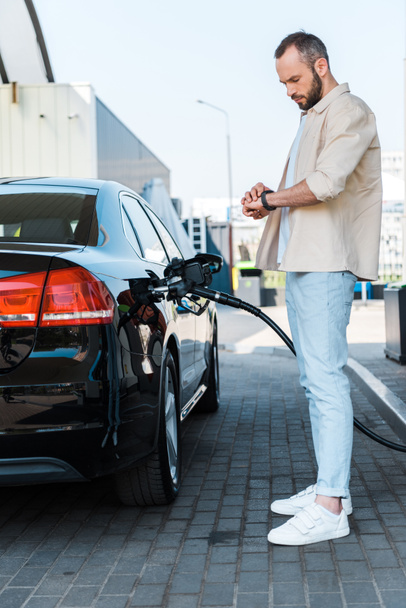 handsome man standing and looking at watch while refueling black car at gas station  - Photo, Image