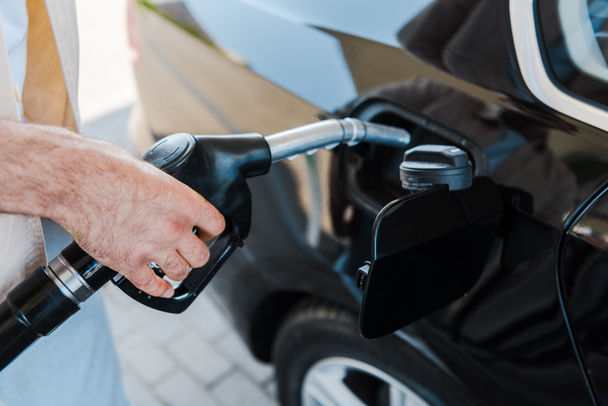 ガソリンスタンドで燃料ポンプを持ち、黒い車に燃料を補給する男のトリミングされた眺め  - 写真・画像