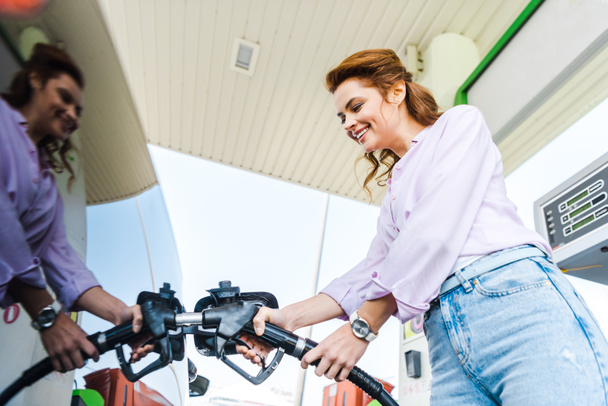 low angle view of happy woman holding fuel pump while refueling car with benzine  - Photo, Image