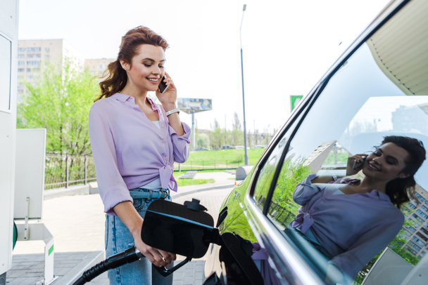 Glückliche Frau hält Benzinpumpe in der Hand, während sie Auto mit Benzin betankt und auf Smartphone spricht  - Foto, Bild