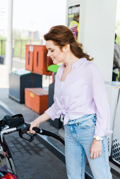 Szczęśliwa kobieta trzyma pompę paliwową podczas tankowania samochodów z benzyna na stacji benzynowej - Zdjęcie, obraz