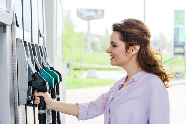 happy woman holding fuel pump while smiling at gas station  - Photo, Image