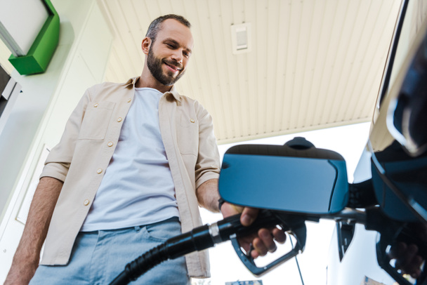 ガソリンスタンドでベンジンで自動車に燃料を補給しながら燃料ポンプを持つ幸せな男の低角度図 - 写真・画像