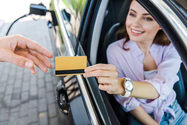 車の中に座って、ガソリンスタンドで労働者にクレジットカードを与える陽気な女性の選択的な焦点  - 写真・画像