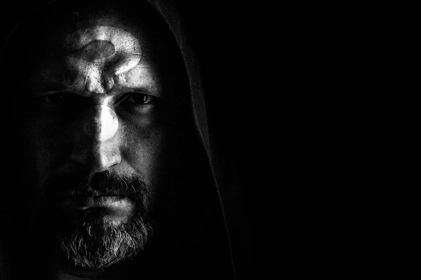 A brutal man with a gray beard, with the question on his face, in a hood with sharp shadows on a black background. Copy space - Photo, Image