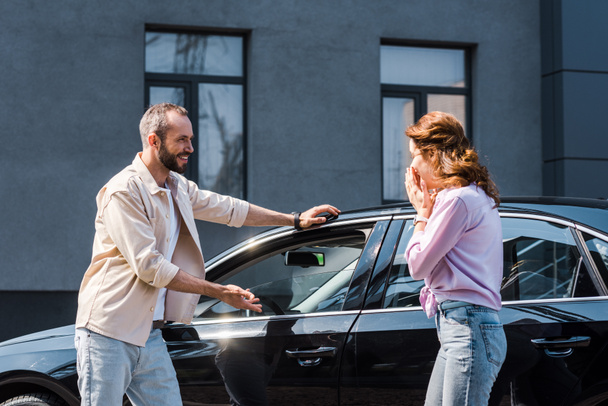 cheerful man gesturing near modern car and standing near excited woman  - Photo, Image