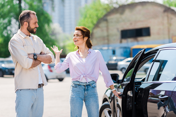szczęśliwy Kobieta wskazując trzymając uchwyt i otwarcie samochodu w pobliżu przystojny mężczyzna  - Zdjęcie, obraz