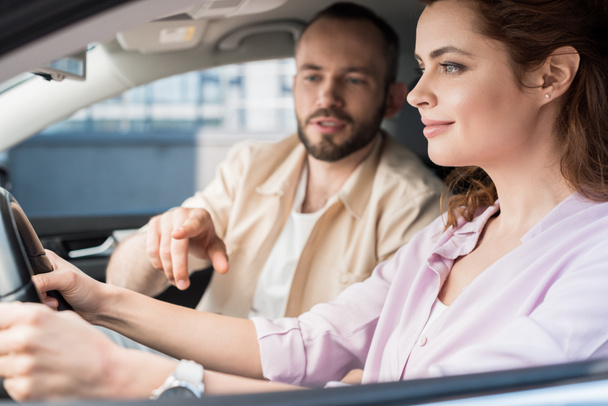 車の中で幸せな女性の近くにステアリングホイールに指で指を指しているハンサムな男の選択的な焦点  - 写真・画像