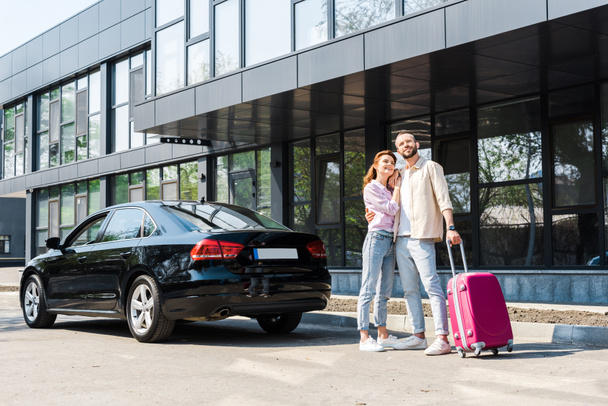 ピンクの荷物を持つ黒い車の近くに立っている間、幸せな女性を抱きしめる陽気な男  - 写真・画像