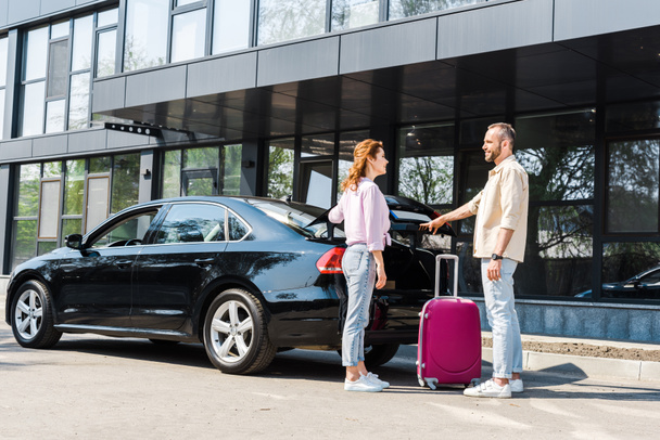 cheerful man looking at happy woman while standing near car and pink luggage  - Photo, Image