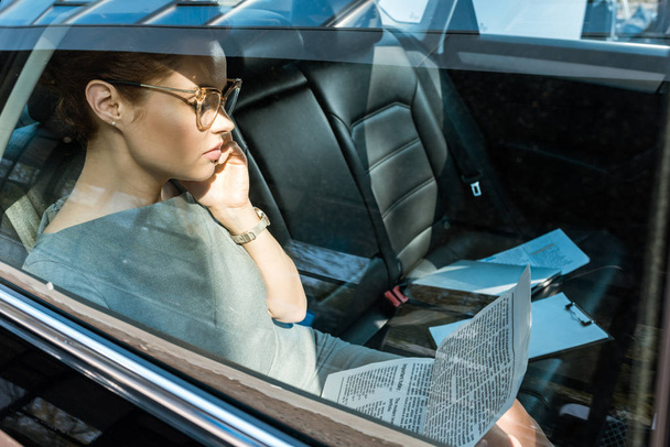 attraktive Geschäftsfrau mit Brille liest Zeitung, während sie im Auto mit dem Smartphone spricht  - Foto, Bild