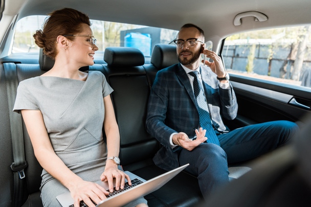 selective focus of businessman in glasses talking on smartphone and looking at businesswoman typing on laptop in car  - Foto, imagen