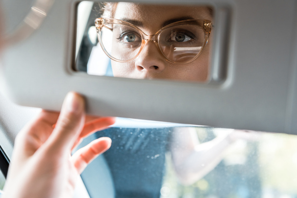 車の中で鏡を見て眼鏡をかけた女性のトリミングされたビュー  - 写真・画像