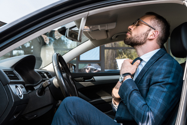 車の中に座っている間にネクタイに触れる眼鏡であごひげのビジネスマンの低角度のビュー  - 写真・画像