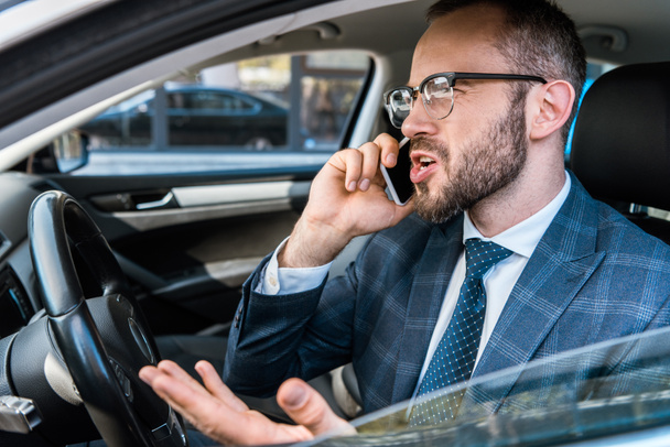 selective focus of angry bearded businessman talking on smartphone and gesturing in car  - Valokuva, kuva