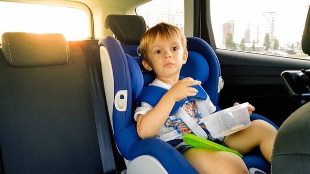 Portrait of 3 years old toddler boy sitting in child safety seat in car and eating cookies. Kids travelling in automobile - Foto, Imagen
