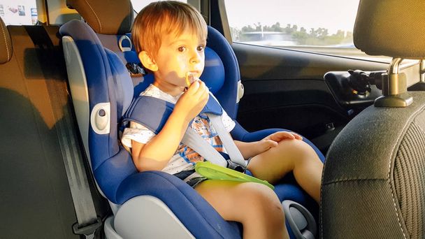 Маленький хлопчик почувається голодним і їсть під час подорожі автомобілем на дитячому сидінні
 - Фото, зображення