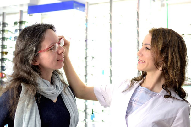 young pretty woman is trying eye glasses on at an eyewear shop with help of a shop assistant, low depth of field - Photo, Image