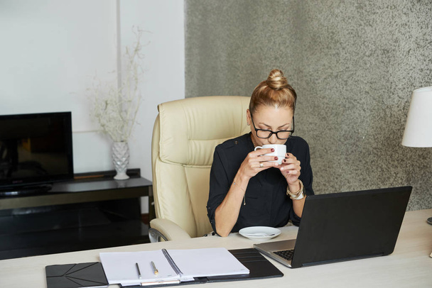 Femme d'affaires dégustant une tasse de café et lisant des e-mails sur l'écran de l'ordinateur portable
 - Photo, image