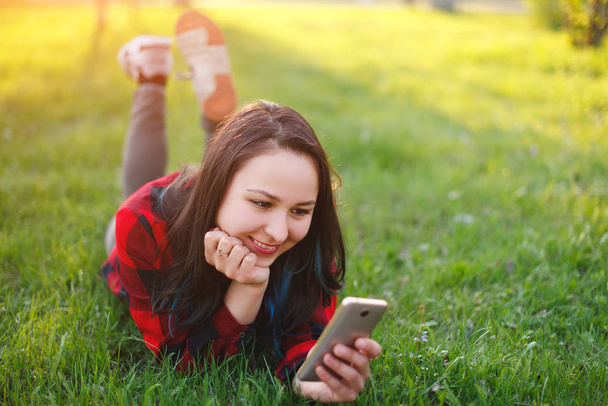 Portrait of a smiling woman lying on green grass and using smartphone outdoors - Фото, изображение