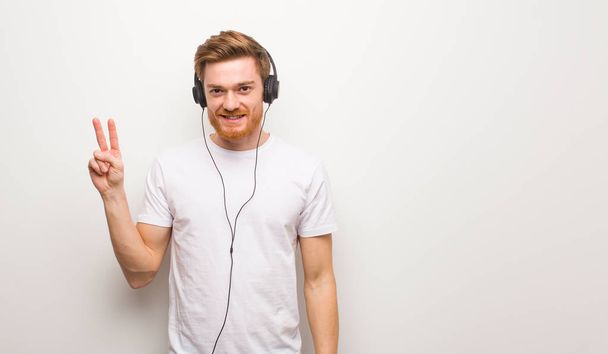 Νεαρός κοκκινομάλλης που δείχνει το νούμερο δύο. Ακούγοντας μουσική με ακουστικά. - Φωτογραφία, εικόνα