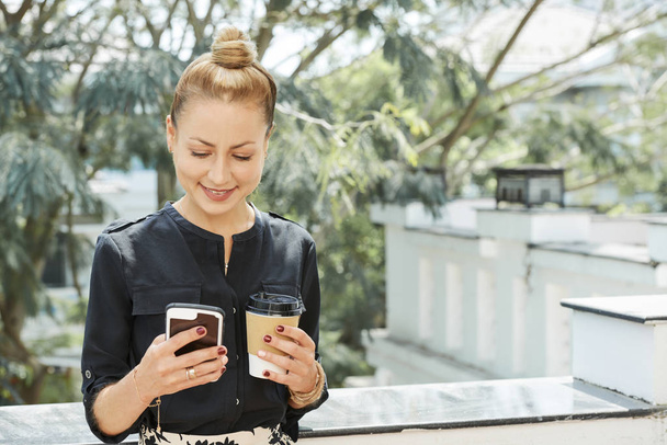 positive hübsche Geschäftsfrau, die draußen mit einer Tasse Kaffee zum Mitnehmen steht und SMS liest - Foto, Bild
