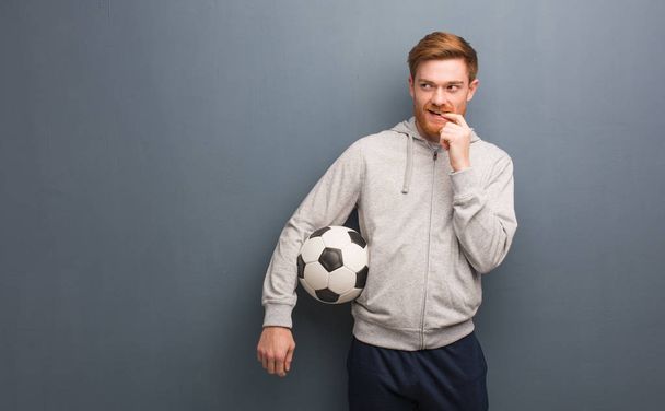Young redhead fitness man relaxed thinking about something looking at a copy space. He is holding a soccer ball. - 写真・画像