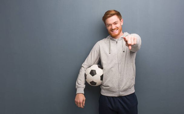 Joven pelirroja fitness hombre alegre y sonriente señalando hacia adelante. Está sosteniendo una pelota de fútbol
. - Foto, Imagen