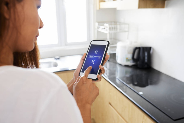 junge Frau mit Smartphone nutzt Home-Service-Anwendung während sie in der heimischen Küche steht - Foto, Bild