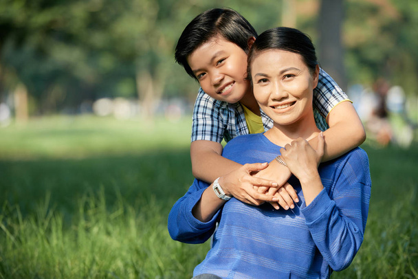 Joyful Vietnamese boy hugging his mother when they are spending weekend in park - Photo, Image