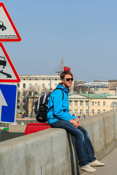 2010.04.11, Moscow, Russia. A young man wearing sunglasses and sitting on the bridge by road sign. - Zdjęcie, obraz