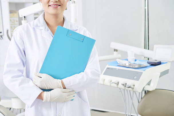 Portrét mladého zubaře, stojícího v uniformě, nosí ochranné rukavice a drží modrou schránku na stomatologické klinice - Fotografie, Obrázek