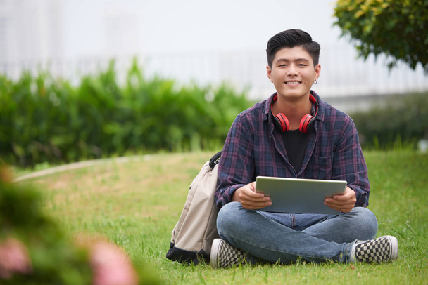 Χαμογελαστός Ασιάτης φοιτητής κολλέγιο με τρυπημένο αυτί κάθεται στο πράσινο γκαζόν με τον υπολογιστή tablet - Φωτογραφία, εικόνα
