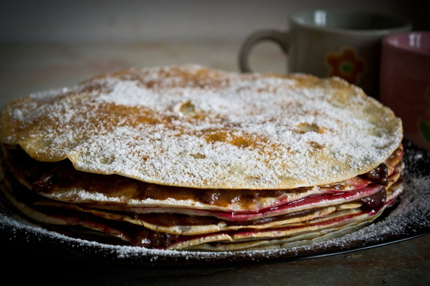 Crepe cake layered with chocolate pudding and jam, sprinkled with powdered sugar with cups in the background - Photo, Image
