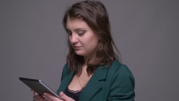Closeup shoot of adult attractive brunette female using the tablet and showing green screen to camera with background isolated on gray - Imágenes, Vídeo