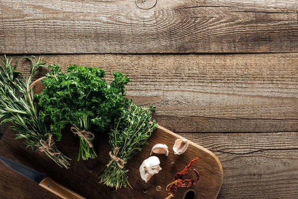 top view of green thyme, parsley, rosemary on wooden chopping board near garlic cloves, knife, dried chili pepper and black pepper on wooden brown table - Zdjęcie, obraz