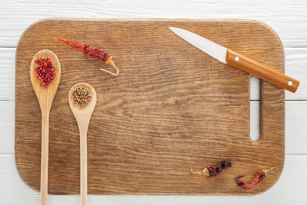 top view of knife, spoons with coriander and pink peppercorn, dried chili peppers on wooden chopping board with copy space - Photo, Image