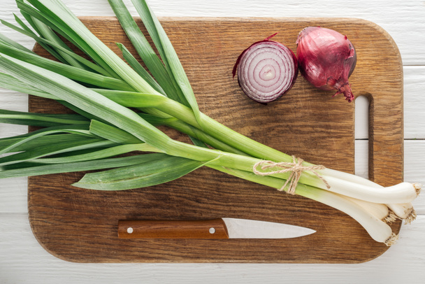 top view of green leek and red onion on wooden chopping board with knife - Фото, зображення