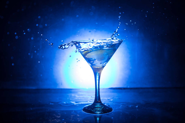 Copa de cóctel Martini en mano salpicando sobre fondo ahumado de tono oscuro o cóctel colorido en vaso con salpicaduras y aceitunas. Entretenimiento del club. Luz mixta. Enfoque selectivo
 - Foto, imagen
