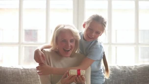 Granddaughter making surprise hugging happy old grandmother presenting birthday gift - Materiał filmowy, wideo